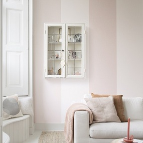 Helles Wohnzimmer mit pastellrosa Wänden, Sofa, Vitrinenschrank und Dekokissen.